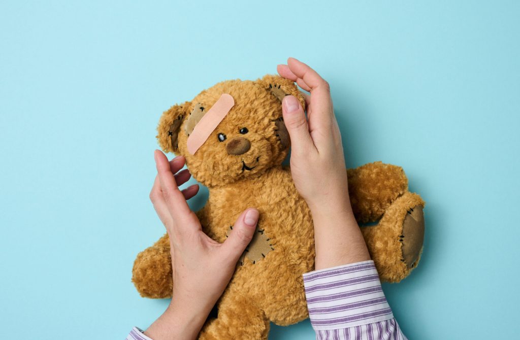 female hand holds a brown teddy bear and glues a medical adhesive plaster on a blue background, tram treatment